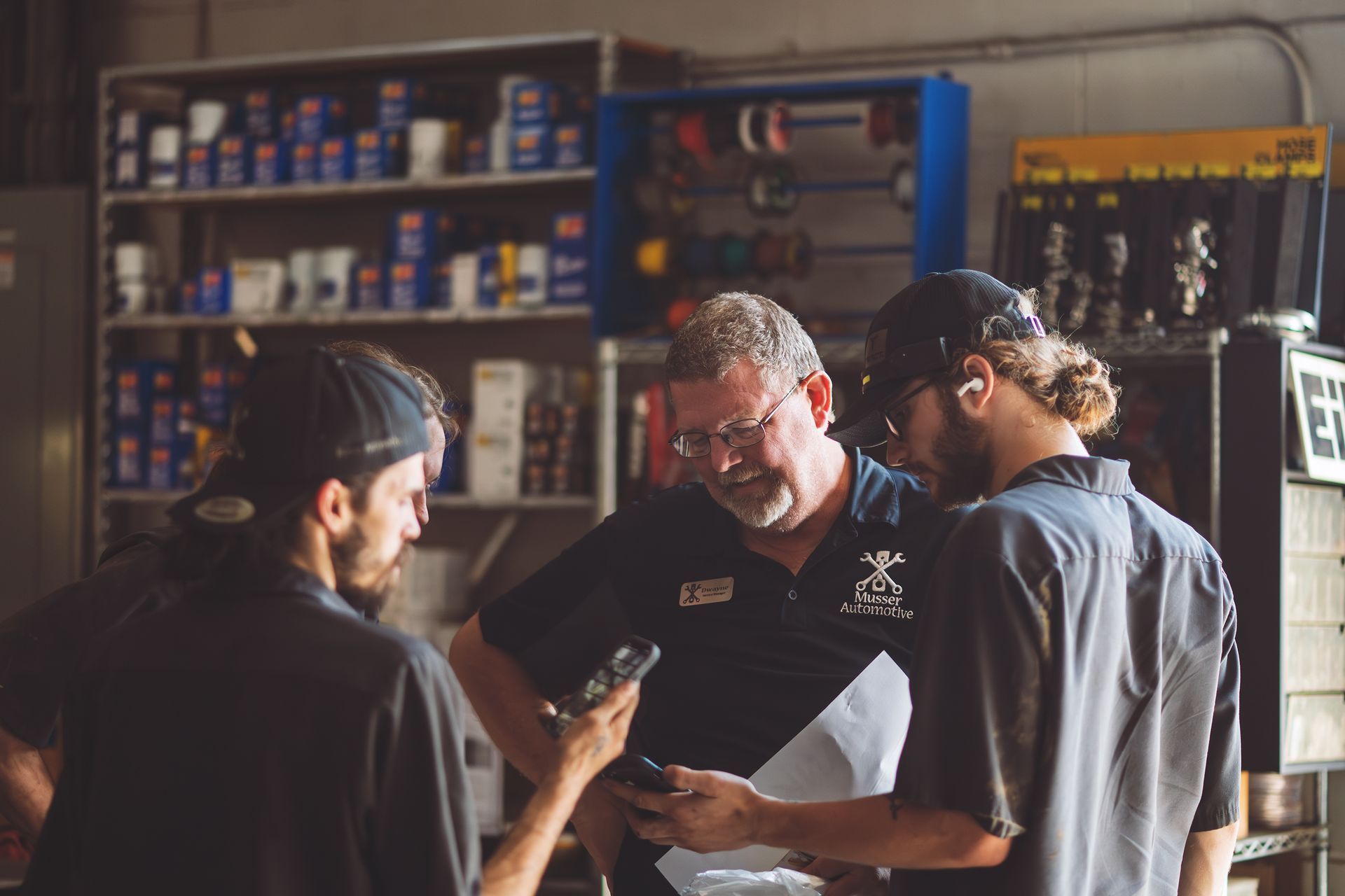 Three mechanics in a shop, reviewing a part and papers. Shelves of supplies in the background. | Musser Automotive