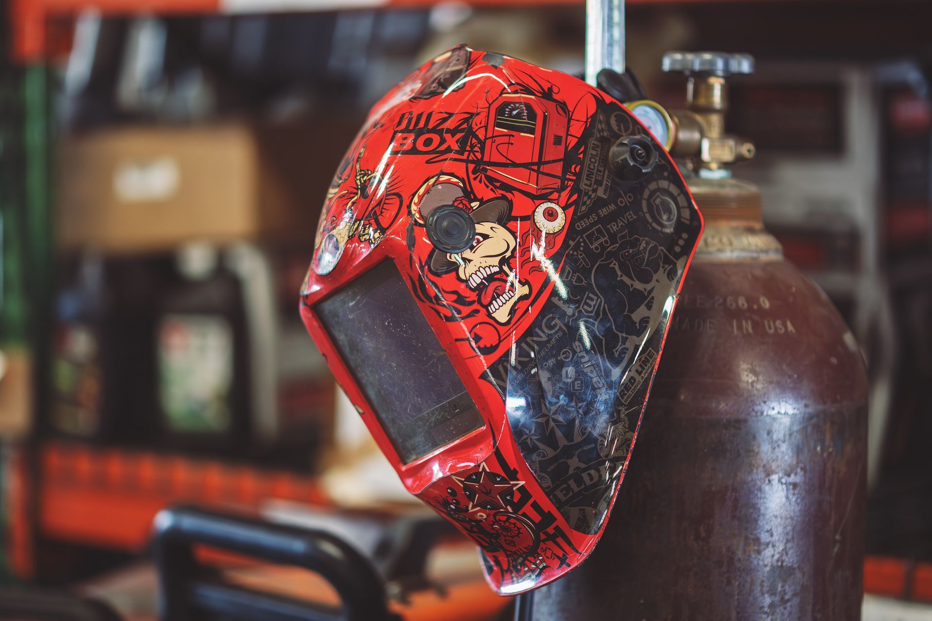 Red welding helmet decorated with skull, hanging next to a brown gas tank. | Musser Automotive