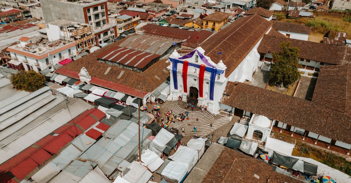 Vista aérea de una plaza con tejados de tejas rojas y un edificio adornado con banderas rojas, blancas y azules.