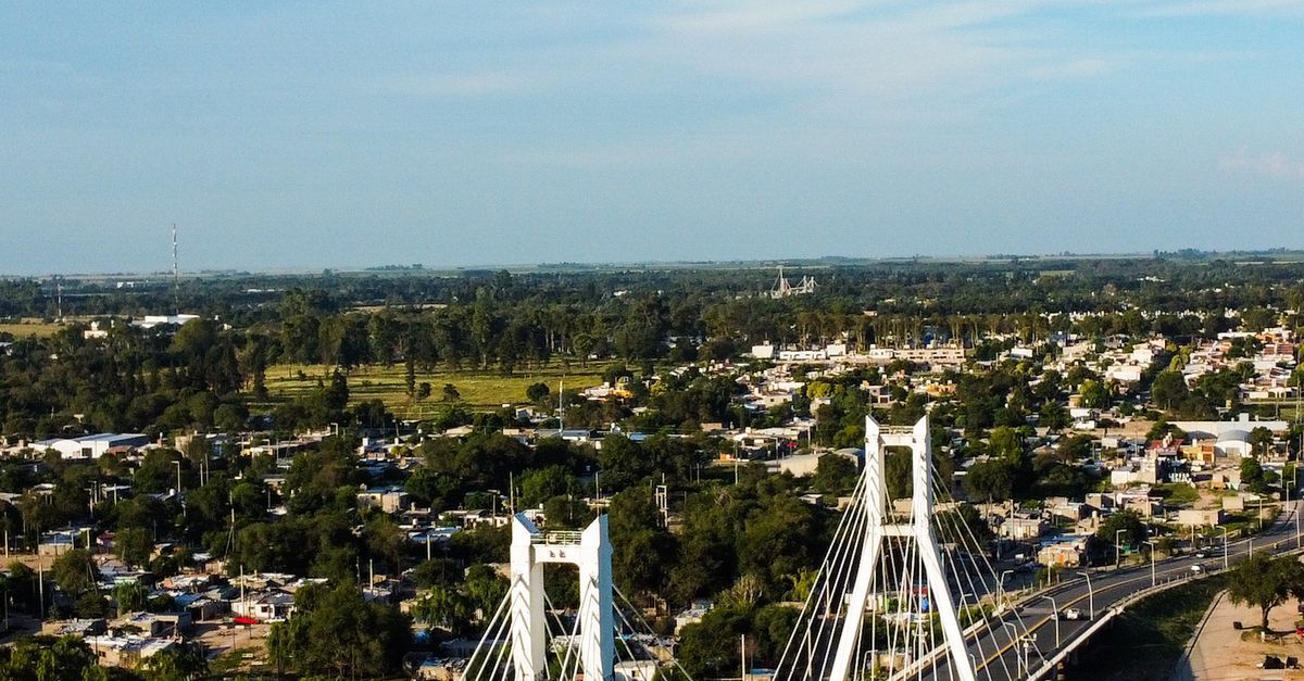 Vista aérea de una ciudad con árboles, barrios y un puente colgante blanco bajo un cielo despejado.