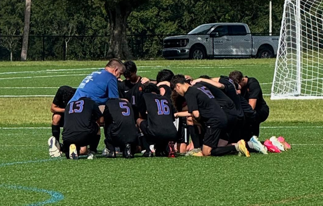 Soccer team huddled kneeling on a green field near a goal, with a pickup truck in the background