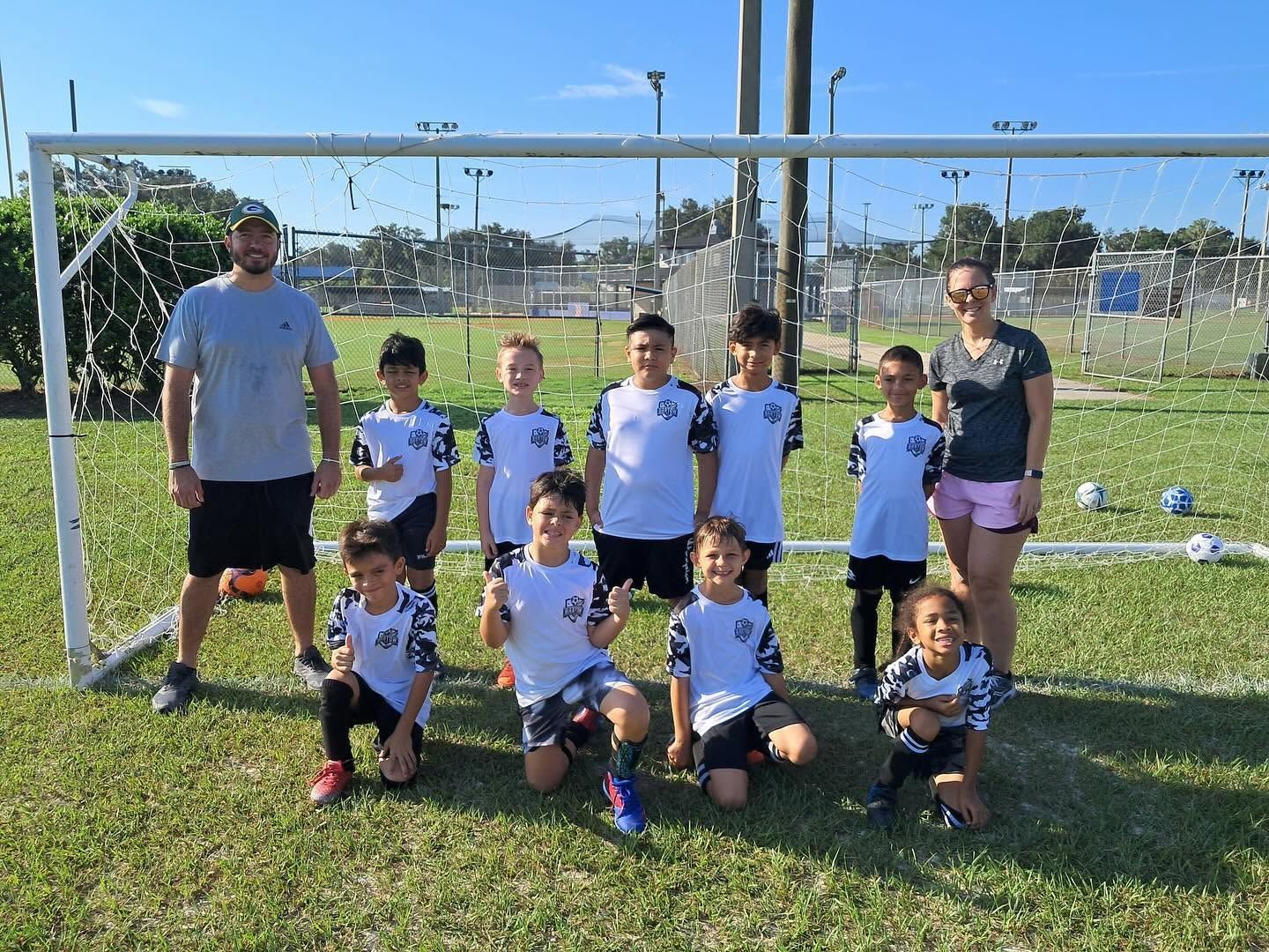 Soccer team posing on a grassy field in front of a goal, with two coaches and children in white uniforms.