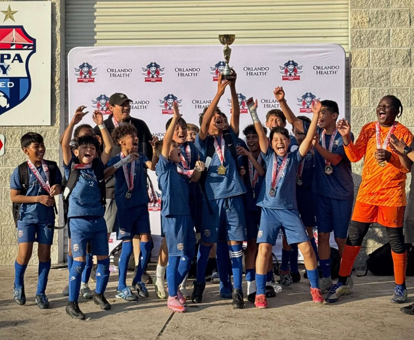 Youth soccer team celebrating with trophy on a field, standing in front of a sponsor backdrop.