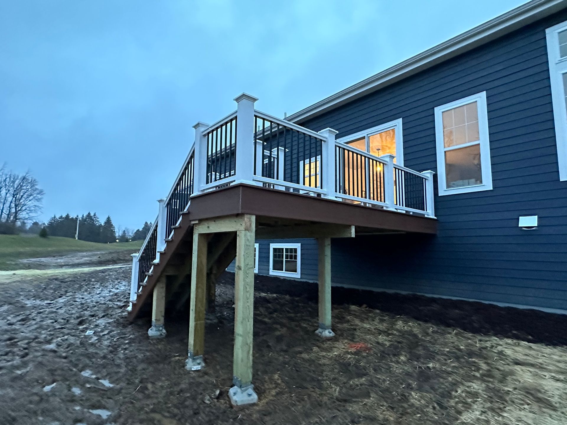 Deck with stairs attached to a dark blue house. White railing and brown decking. Cloudy sky.