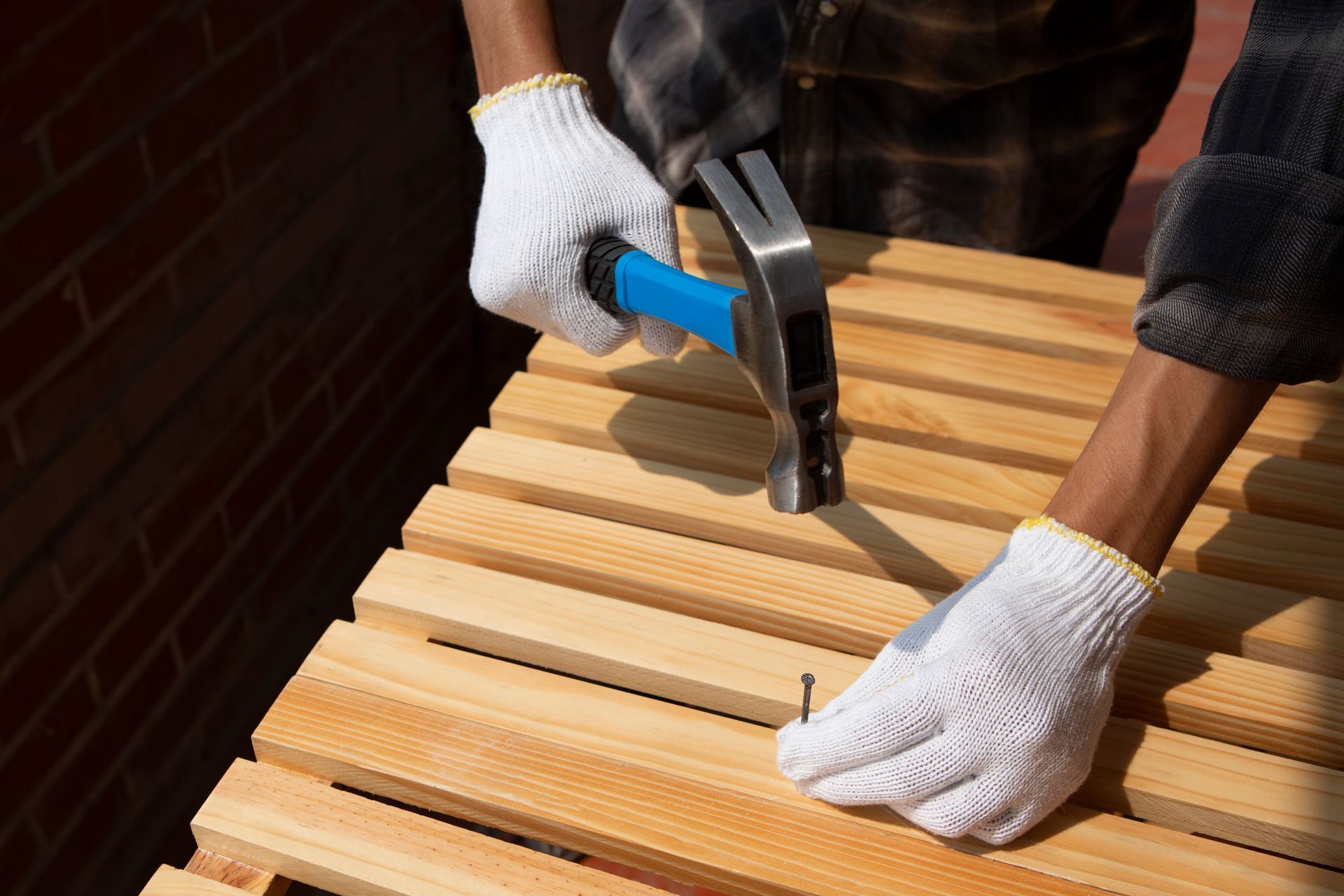 Gloved hands hammering nails into a light wooden slatted bench outdoors