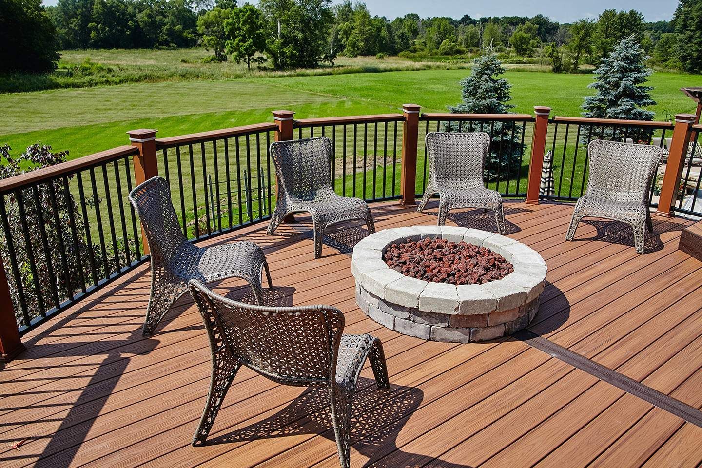 Wooden deck with six chairs arranged around a stone fire pit overlooking a green field.