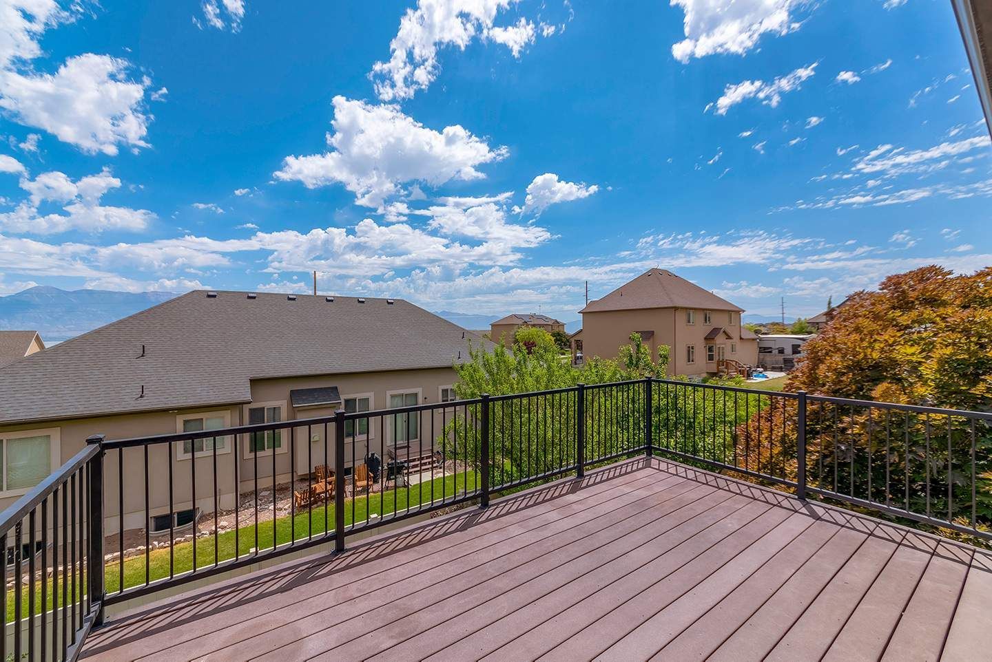 View from a deck with brown decking, black railing, towards houses, trees, and a blue sky with clouds.