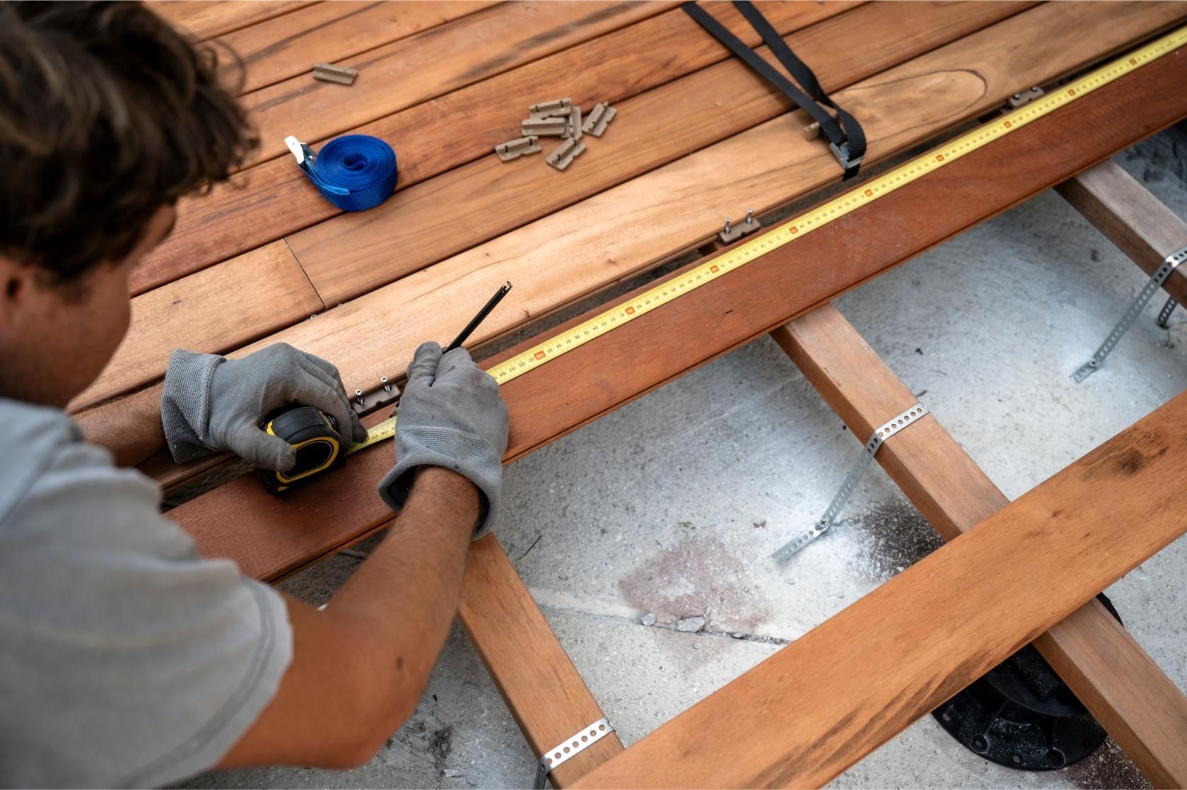 A person wearing work gloves uses a pencil and tape measure to mark a wooden deck plank during construction.