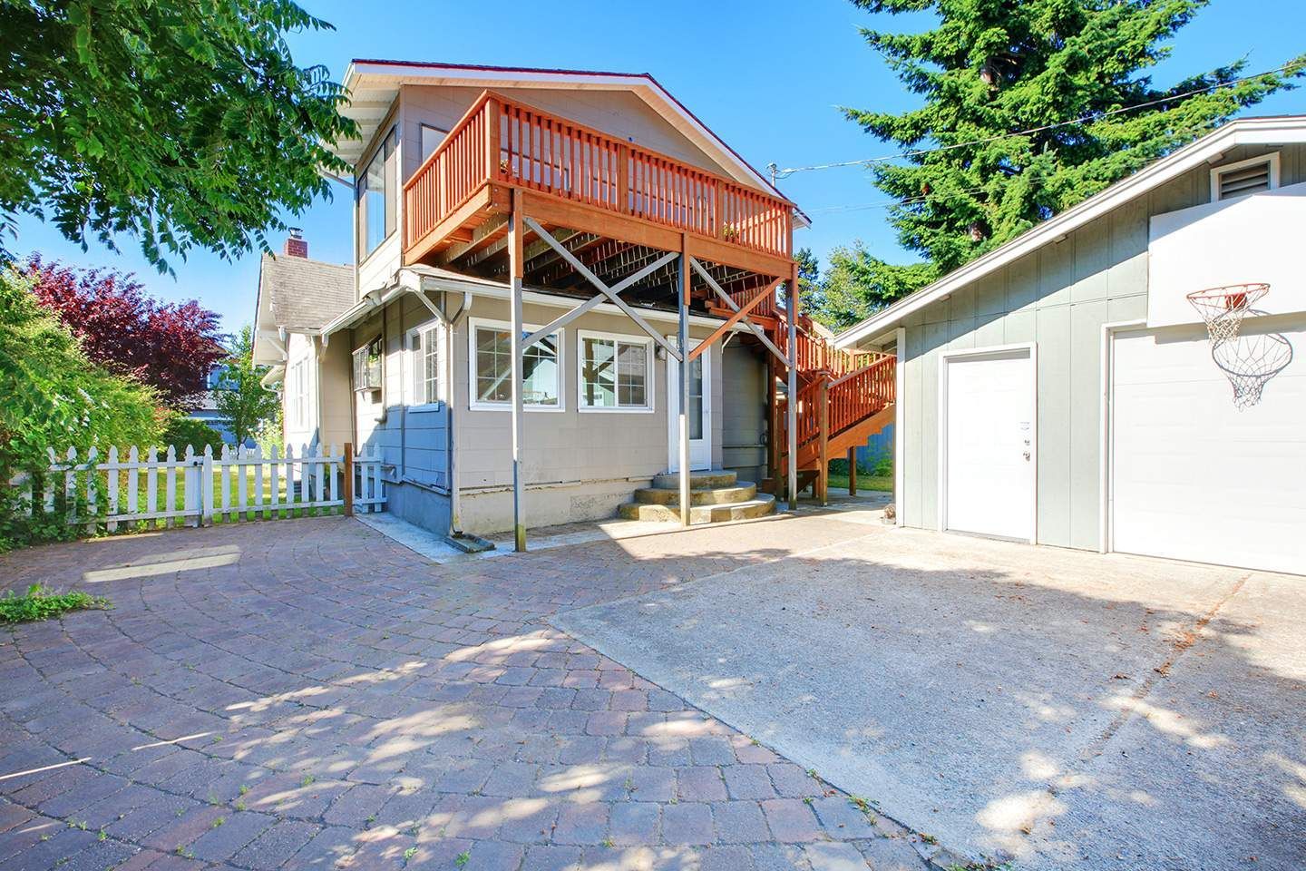 House with a second-story deck, driveway, white picket fence, and detached garage with a basketball hoop.