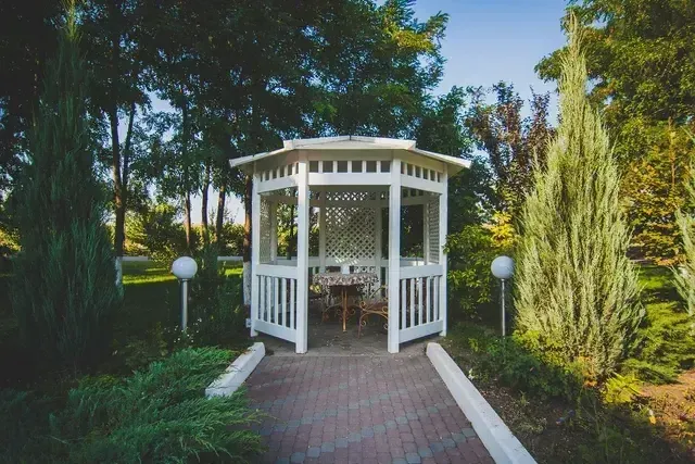 White gazebo on a brick path, surrounded by greenery and trees in a sunny park setting.