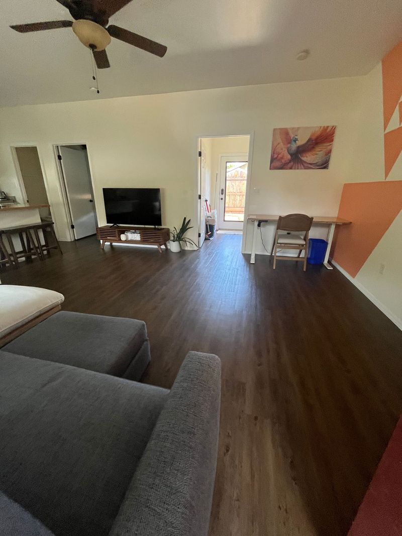 Open living room with gray sofa, TV, ceiling fan, wood floors, and orange accent wall