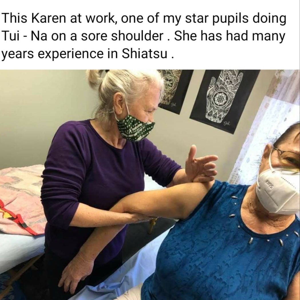 Two women, one in purple, massages the sore shoulder of another in blue, both wearing masks.
