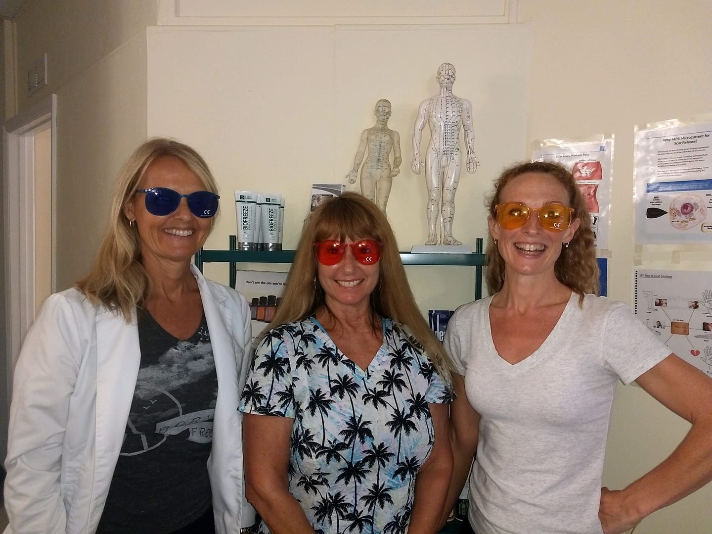 Three women smiling, wearing oversized sunglasses in blue, red, and yellow, standing indoors near anatomical models.