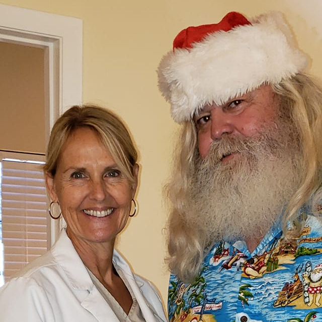 Woman in white coat smiles next to a man dressed as Santa in a Hawaiian shirt.