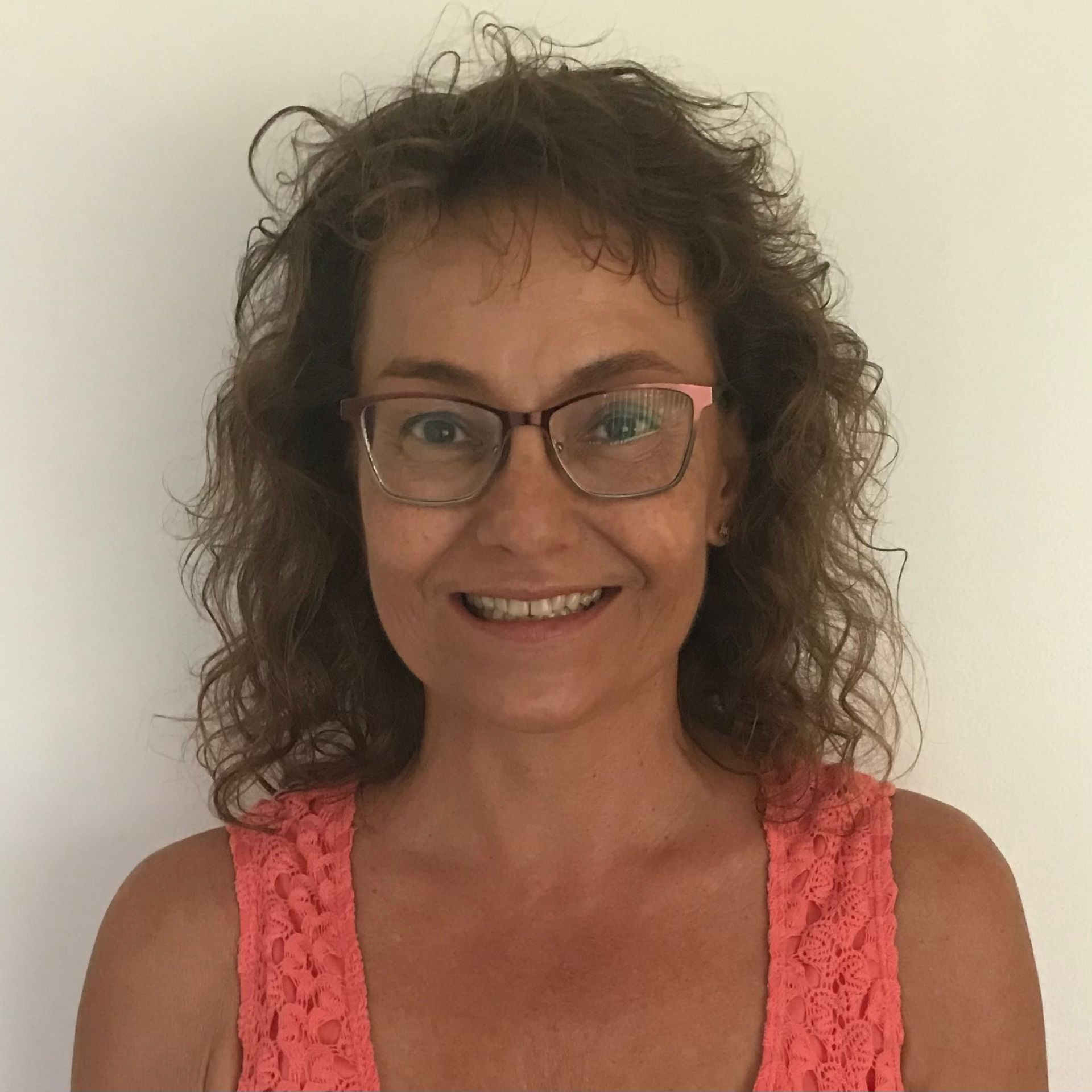 Woman with glasses smiles at the camera, wearing a coral top against a white wall.