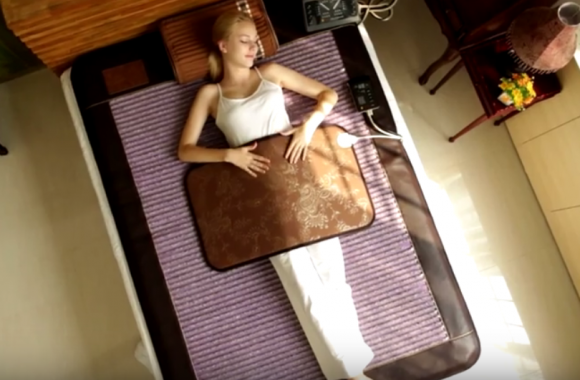 Woman lying on a bed with a heating mat, sunlit room.