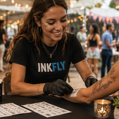 Tattoo artist in black gloves applies a stencil to a client’s forearm at an outdoor event.