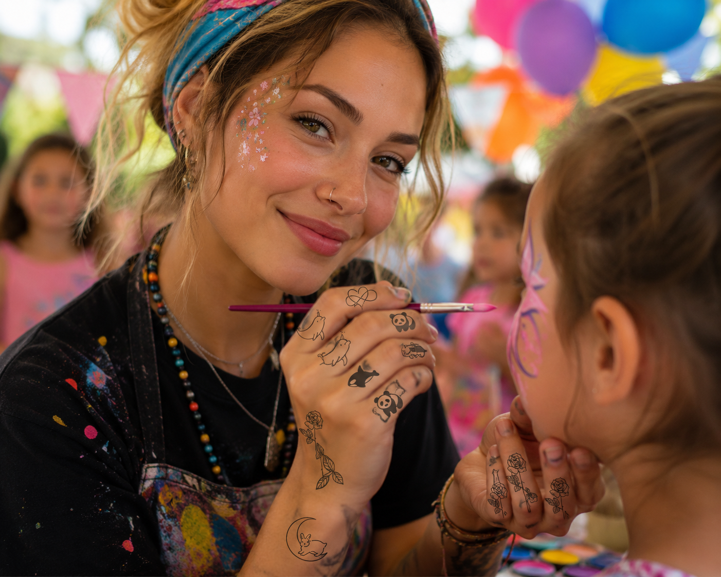 Person with face paint smiles while painting a child’s face at a colorful party with balloons.