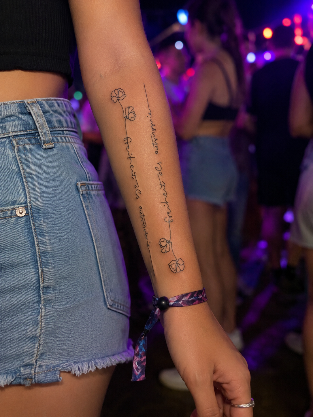 Denim shorts and a black top in a neon-lit nightclub, showing a floral arm tattoo.