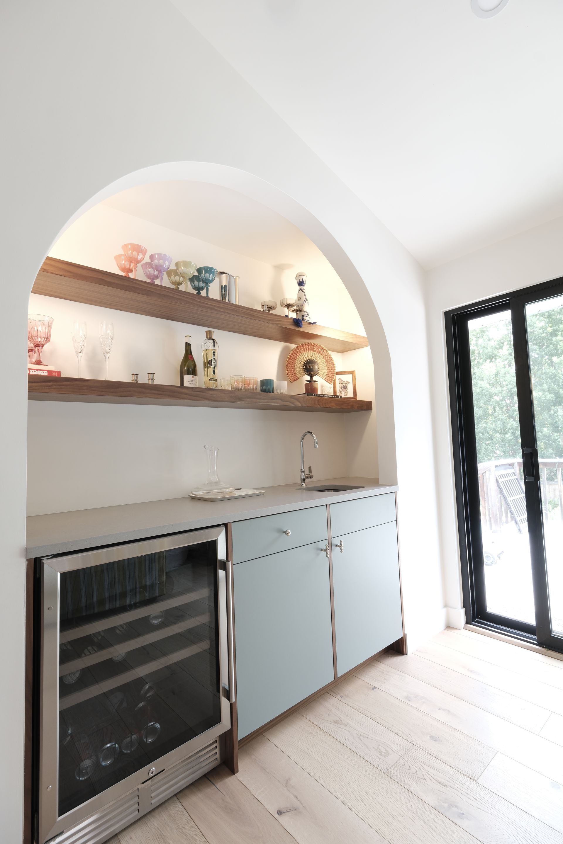 Bright kitchen nook with arched shelves, mini fridge, pale cabinets, and a black-framed glass door