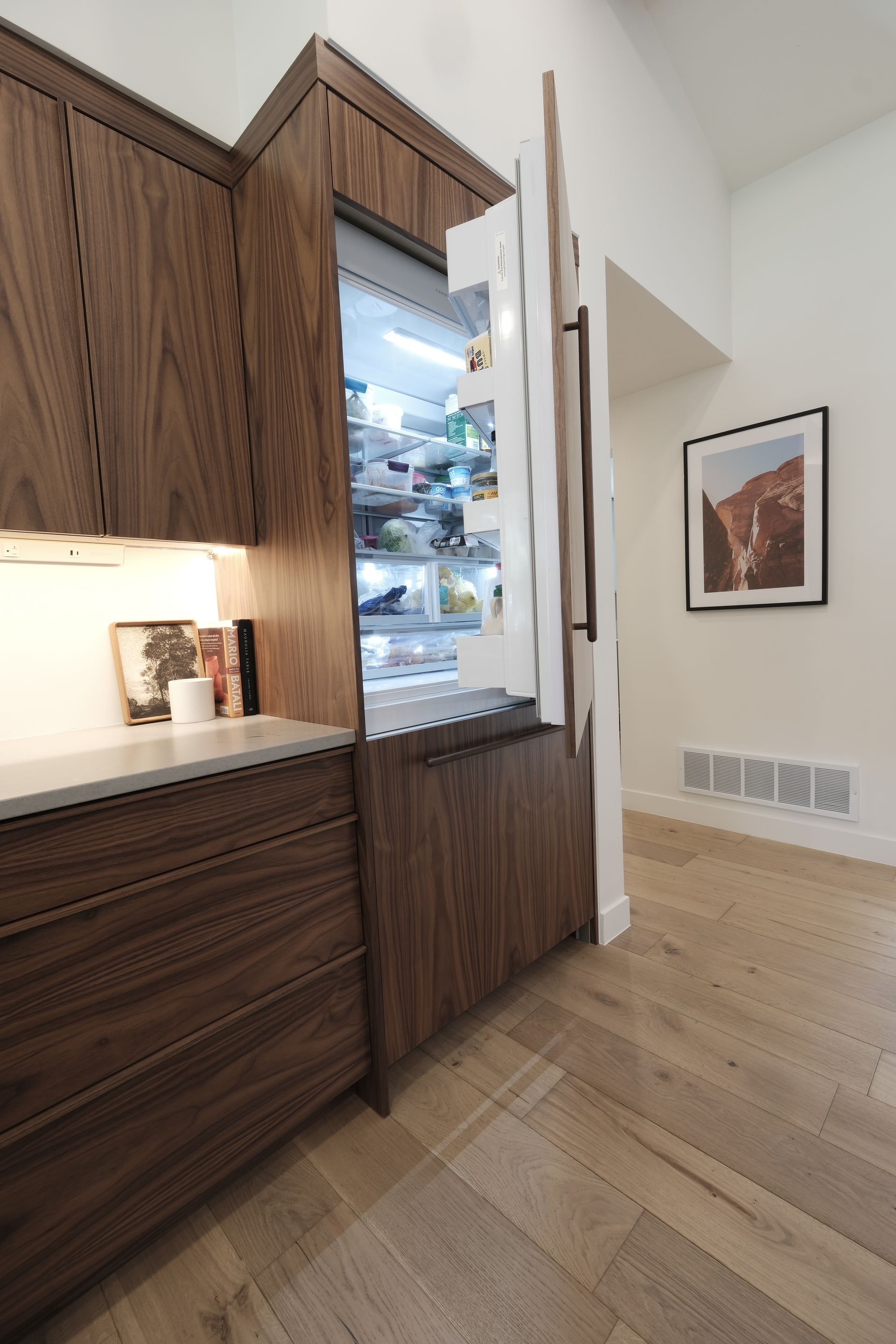 Modern kitchen with wood cabinets, open refrigerator, and framed wall art on a light wood floor