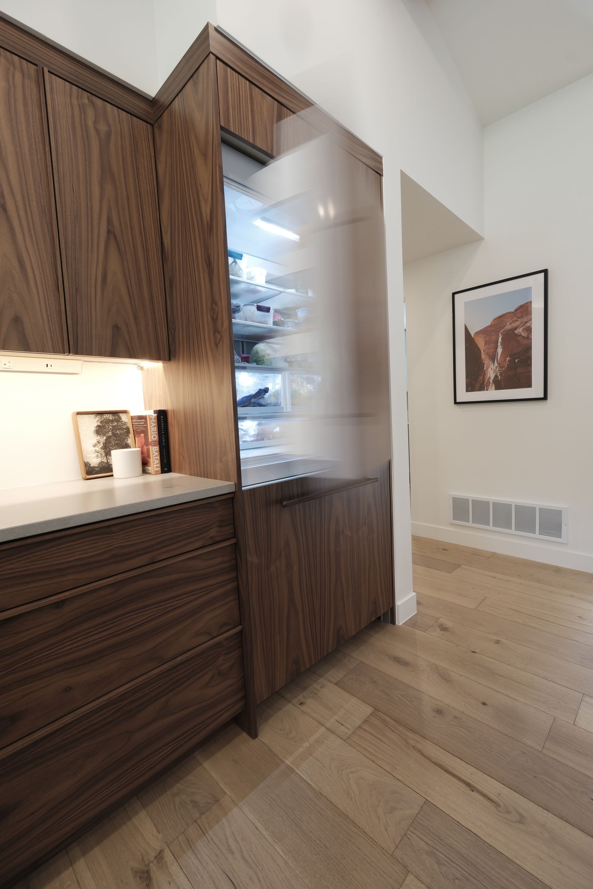 Modern kitchen with wood cabinets, built-in refrigerator, and hardwood floor beside a framed wall print