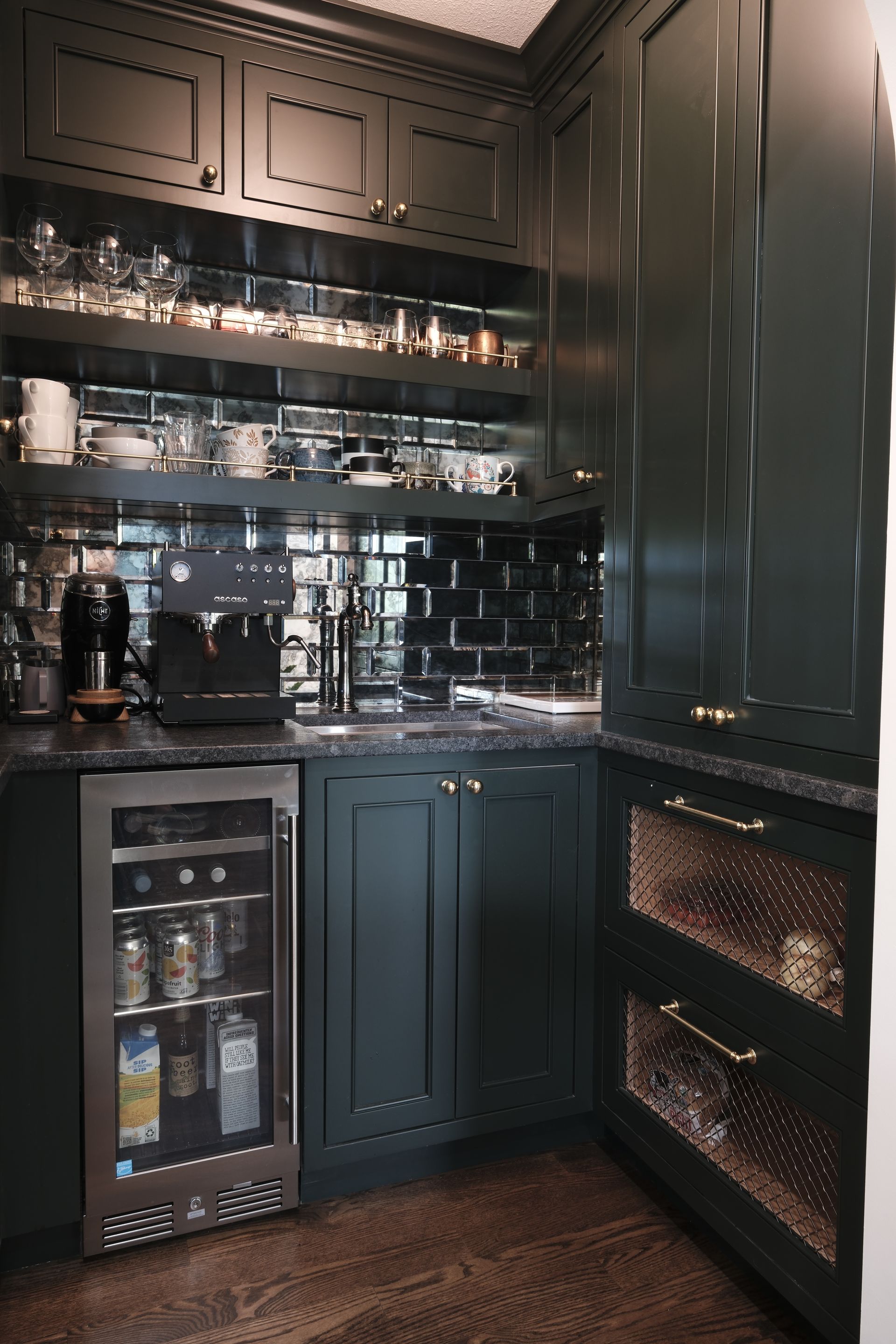 Dark, elegant kitchen nook with shelves of glassware, black cabinetry, and a wine fridge under warm lighting