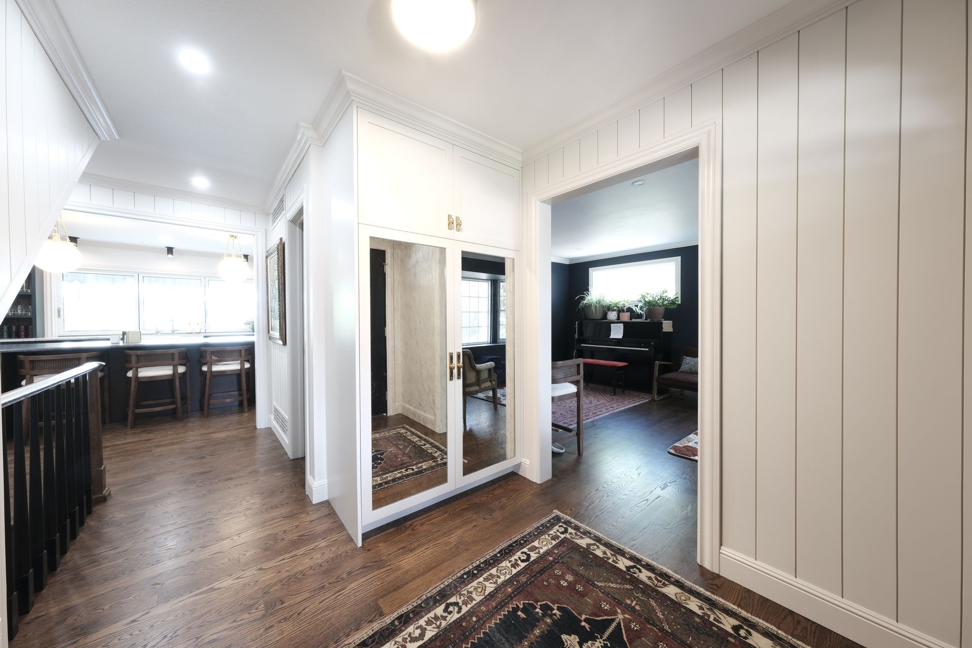 Bright hallway with white walls, wood floors, and a mirrored closet opening into a dark room and kitchen area.