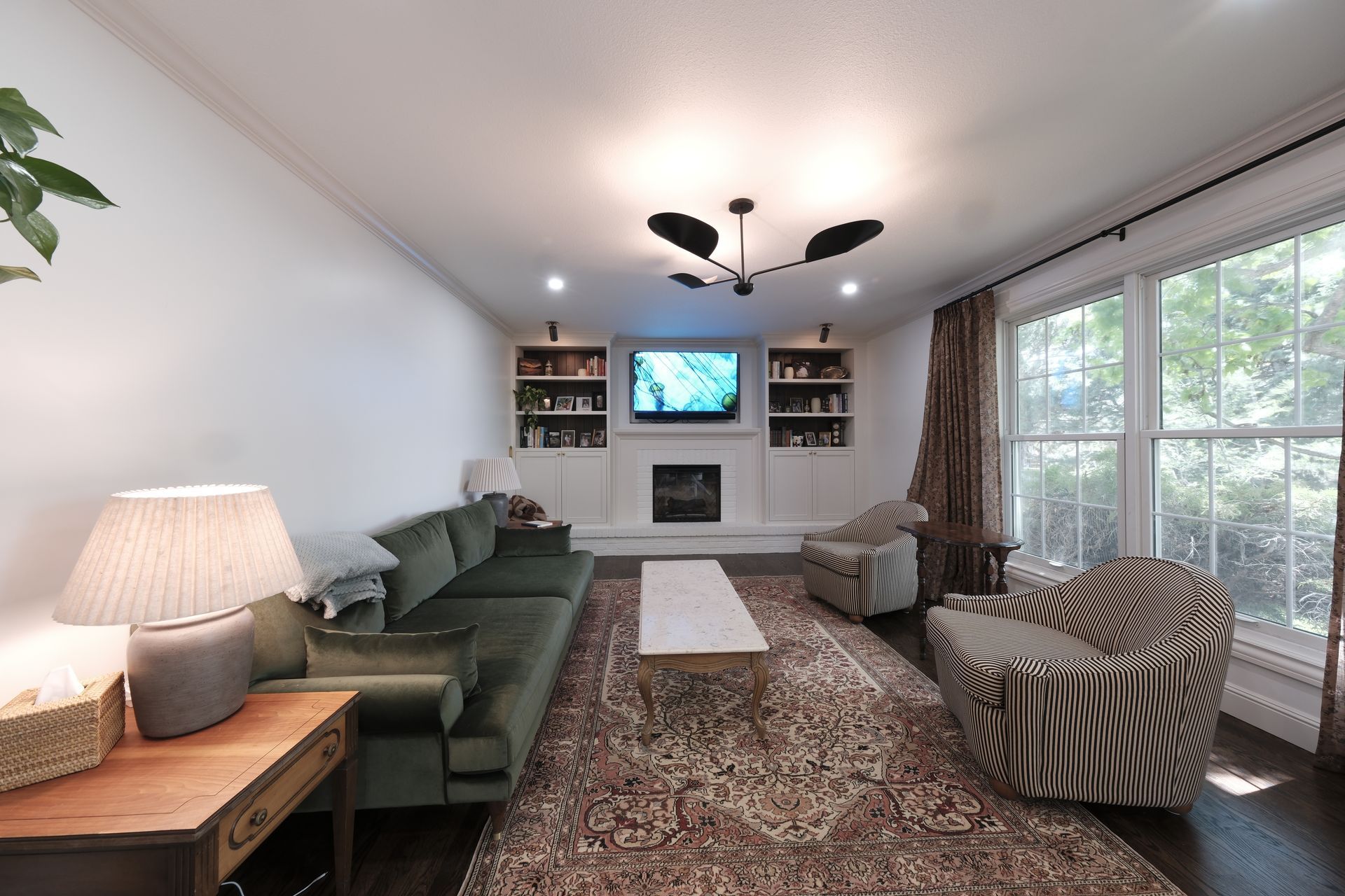 Bright living room with green sofa, patterned chairs, large windows, rug, and a TV above a fireplace.
