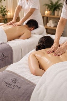 Two people receive back massages on white towels in a dim spa with lit candles.