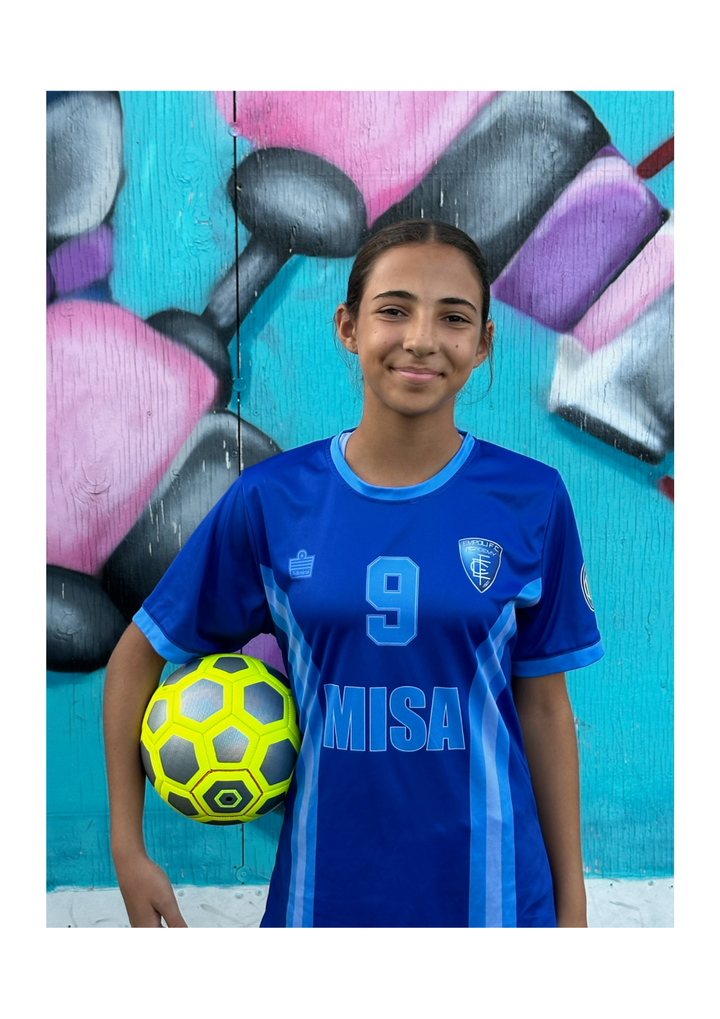 A girl in a blue jersey is holding a soccer ball in front of a graffiti wall.