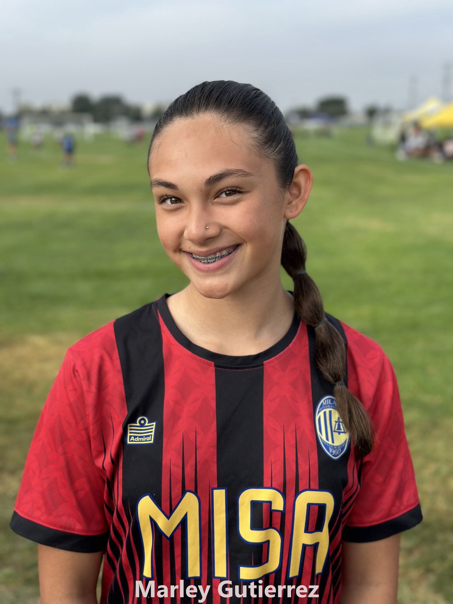 Soccer player, Marley Gutierrez, in red and black jersey smiles at the camera on a grassy field.