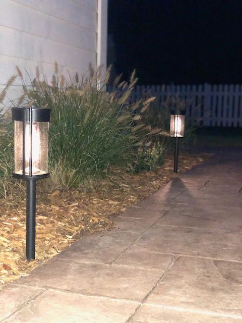 Solar path lights illuminate a stone walkway beside a house and shrubbery at night.