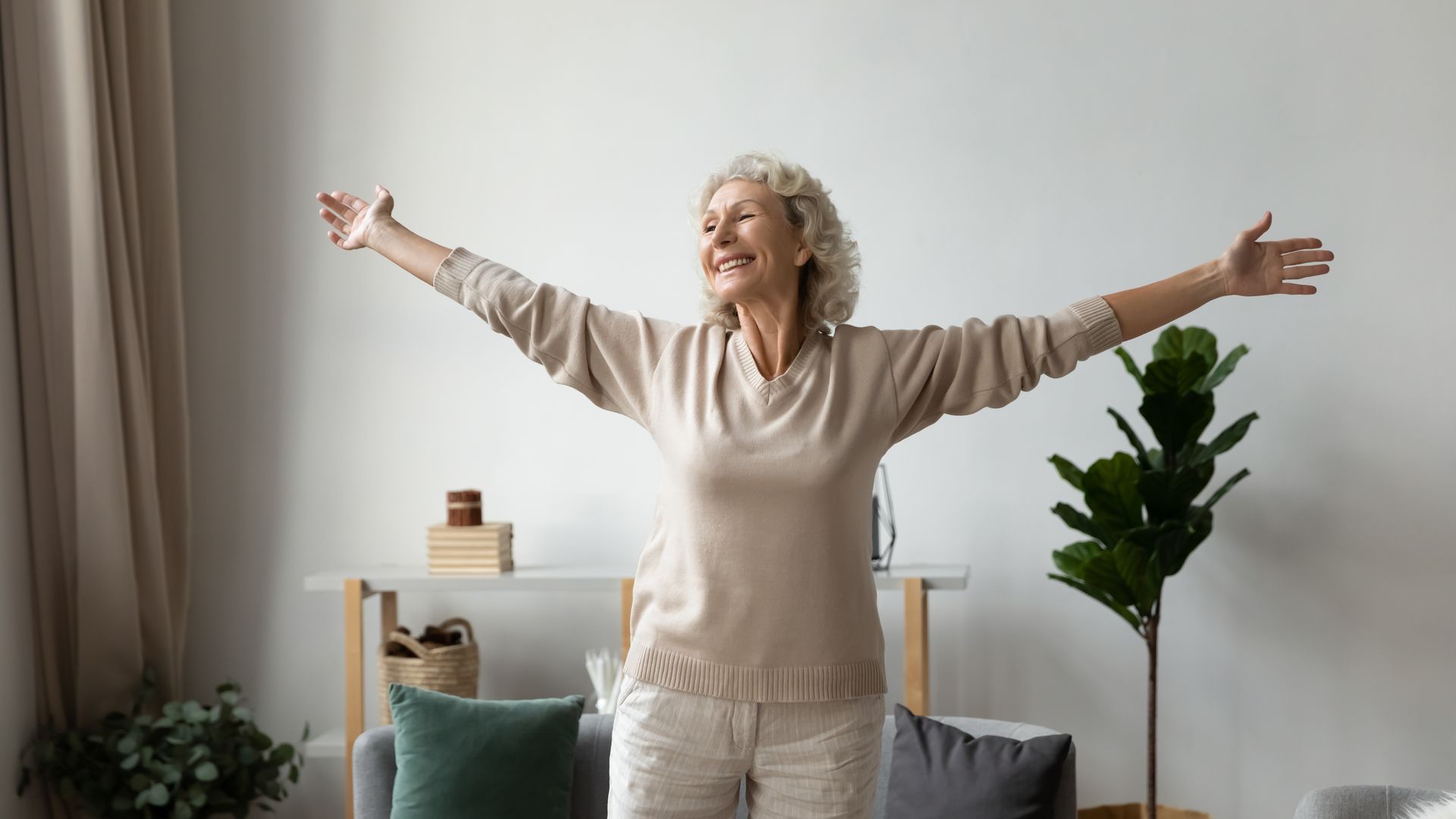 Smiling older woman with arms outstretched in a light-filled living room, appearing happy and content.