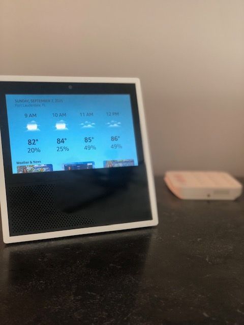White smart display showing weather forecast, sitting on a black surface next to a small white device.