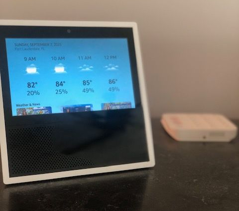 An Amazon Echo Show displays the weather forecast. A small white device is on a table.