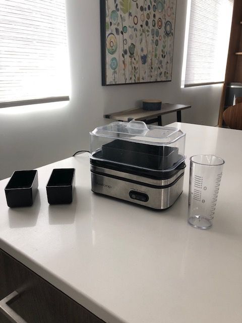Food steamer on a white counter with two black containers and a measuring cup.