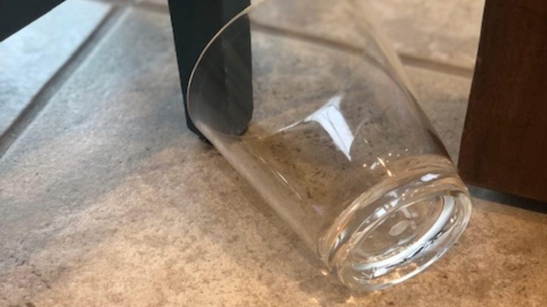 A clear glass on its side on a tile floor, near a dark chair leg and brown furniture.