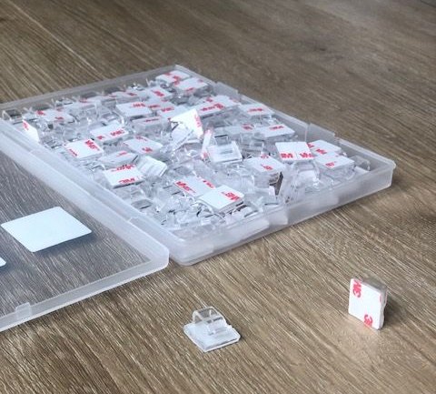 A clear plastic case holding many small cable clips with adhesive pads. Some clips are outside the case on a wood surface.