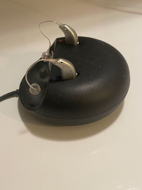 Two silver hearing aids in a black charging base on a white surface.