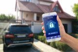 Hand holding a smartphone displaying a home security app with a house and car in the background.