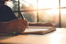 Close-up of a person writing in a notebook with a pen at a wooden table near a sunlit window.