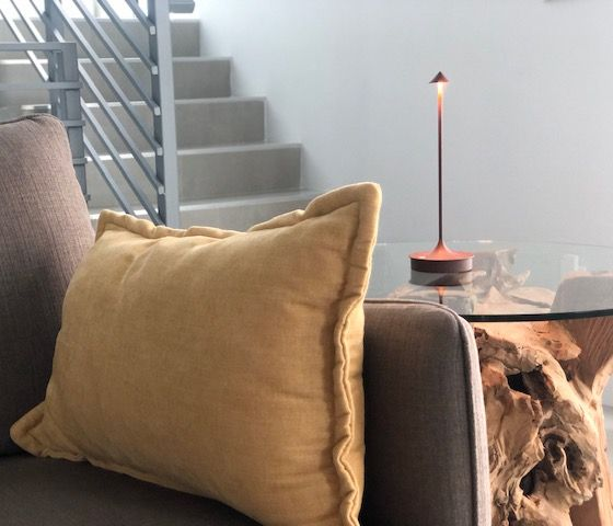 Yellow pillow on gray sofa with a small lamp on a glass table in front of stairs.