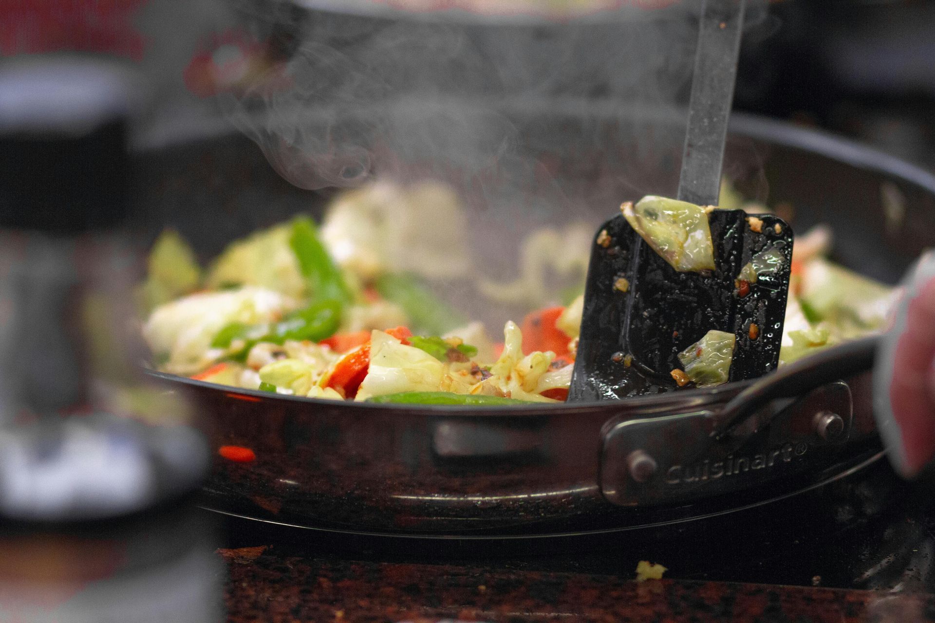 Vegetables cooking in a pan; steam rising, spatula in use.