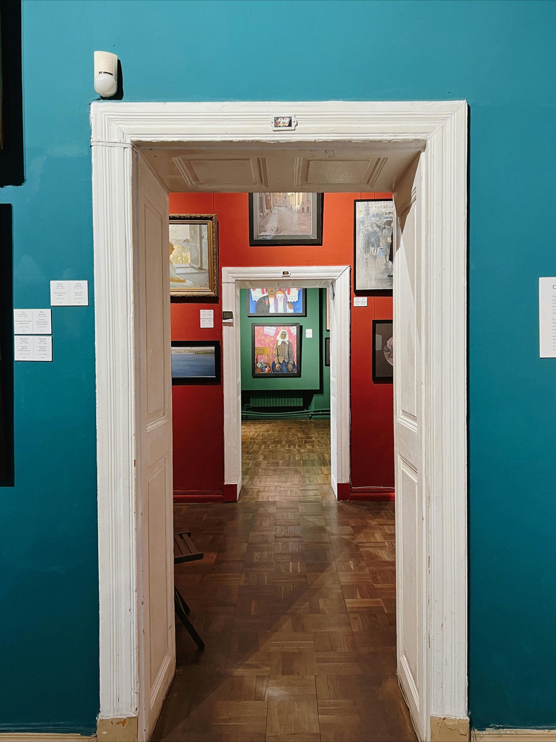 View through doorways into a gallery with paintings on red and green walls. White door frames contrast with teal walls.