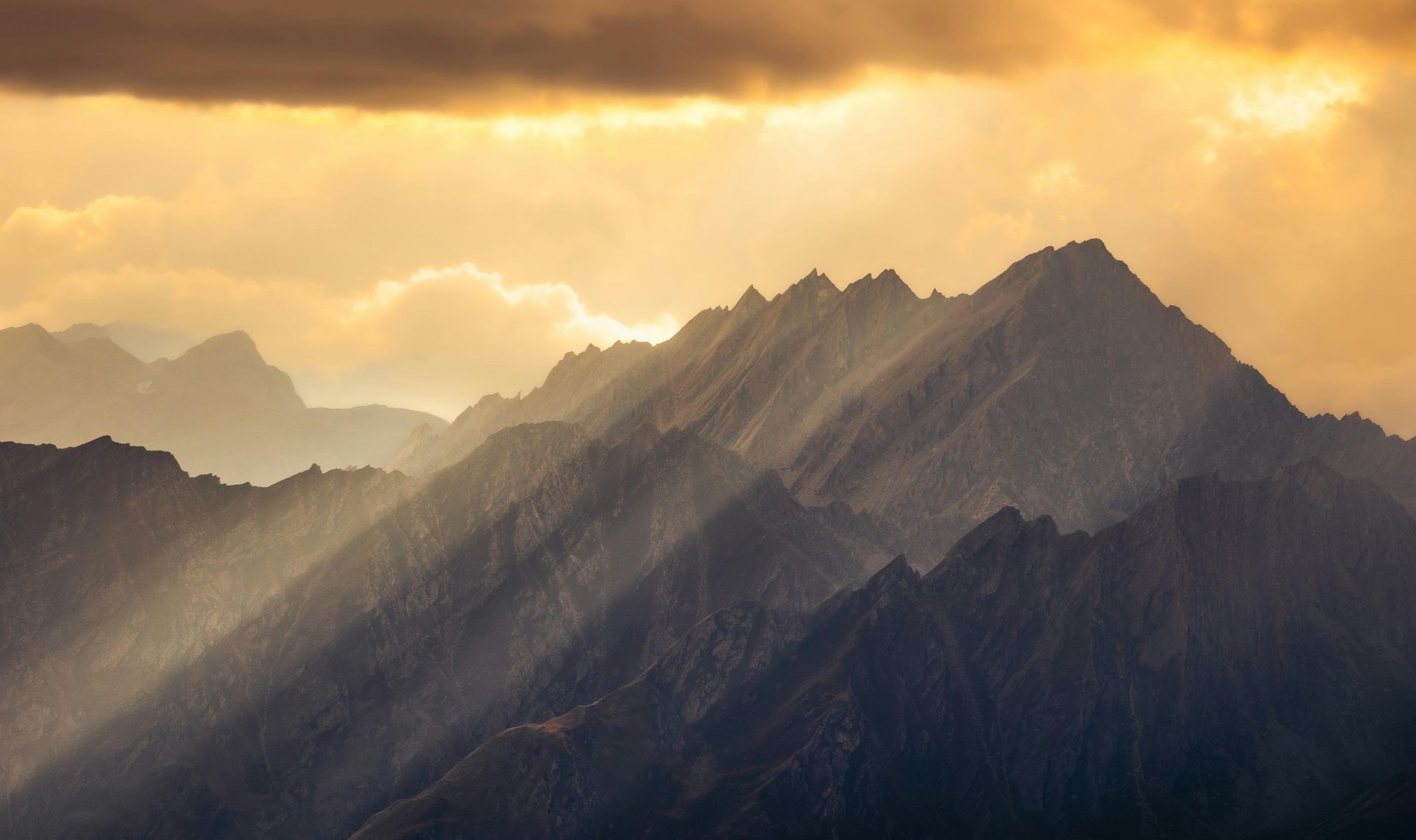 Sunlight streams over jagged mountain peaks through a cloudy sky; golden light.