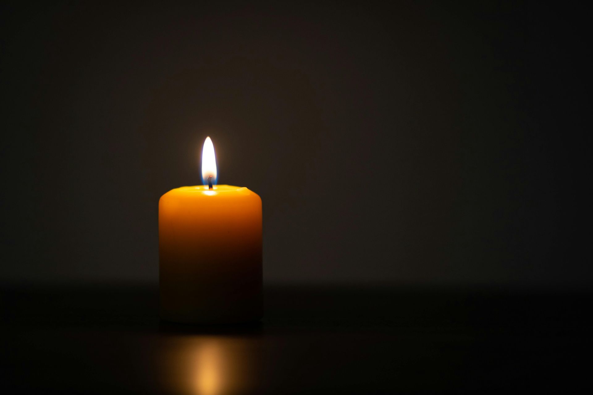 A lit yellow candle on a dark surface, casting a warm glow with a dark background.