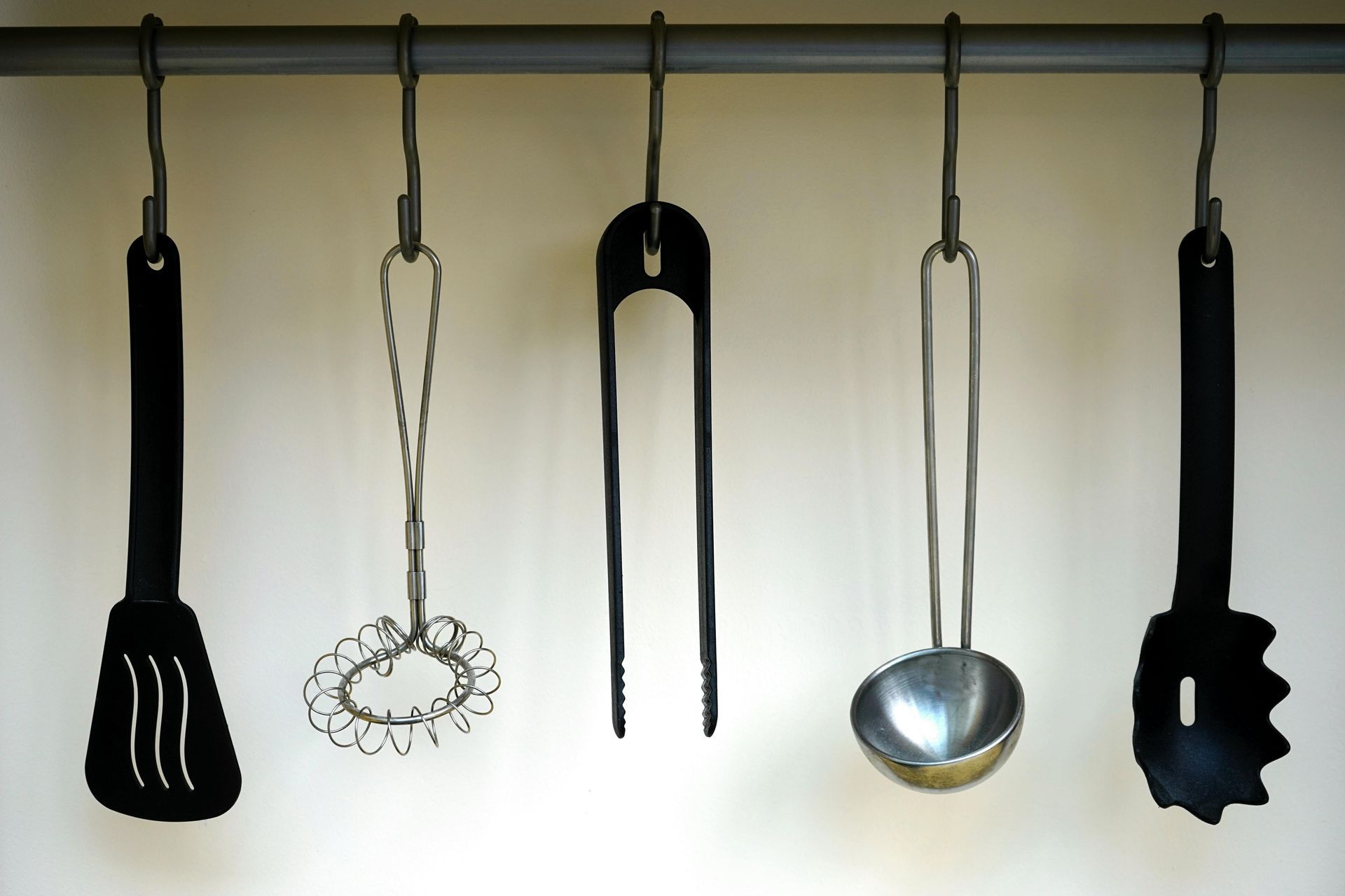 Kitchen utensils hanging on a metal bar: spatula, whisk, tongs, ladle, and pasta server.