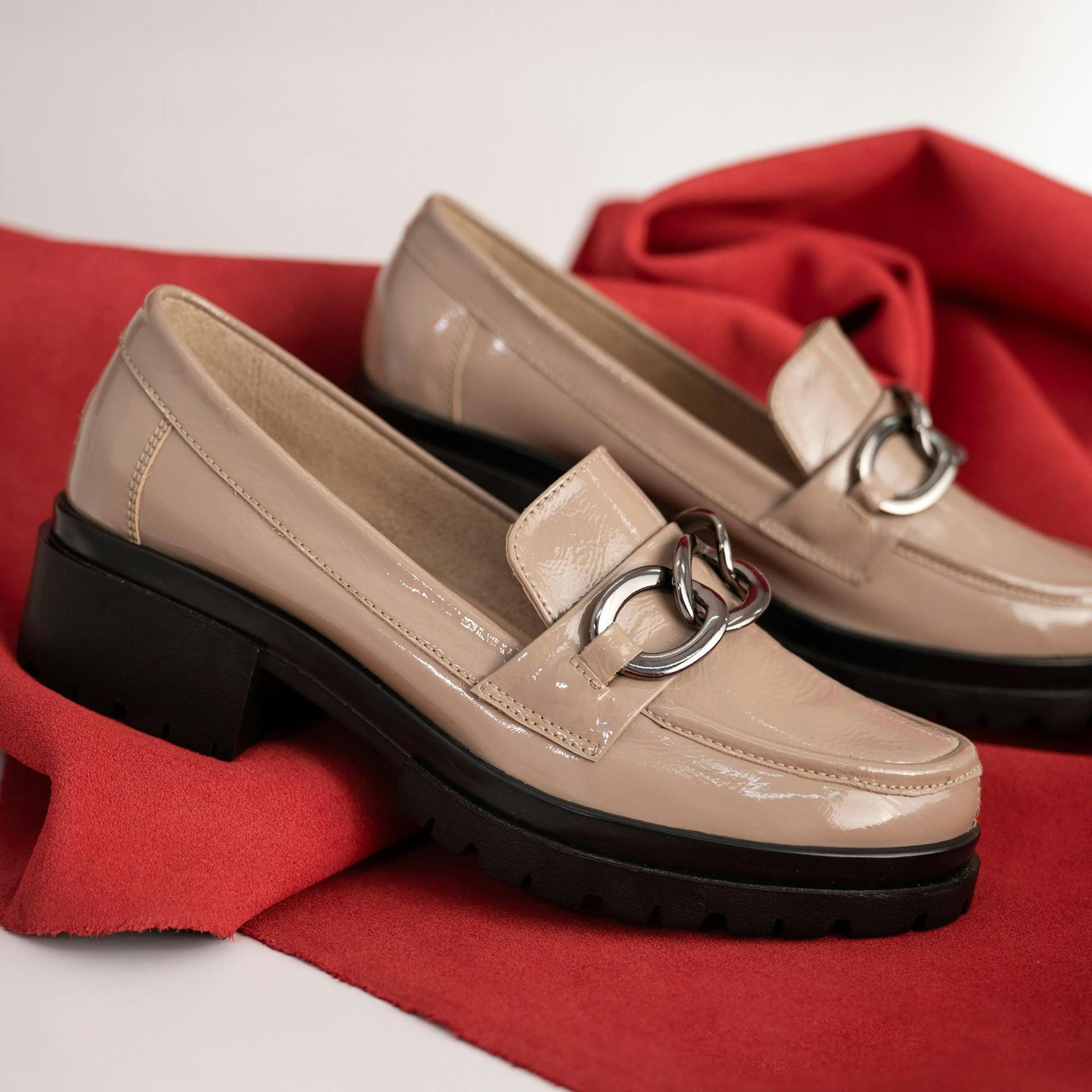 Beige patent leather loafers with silver chain detail, set on a black platform sole, resting on red fabric.