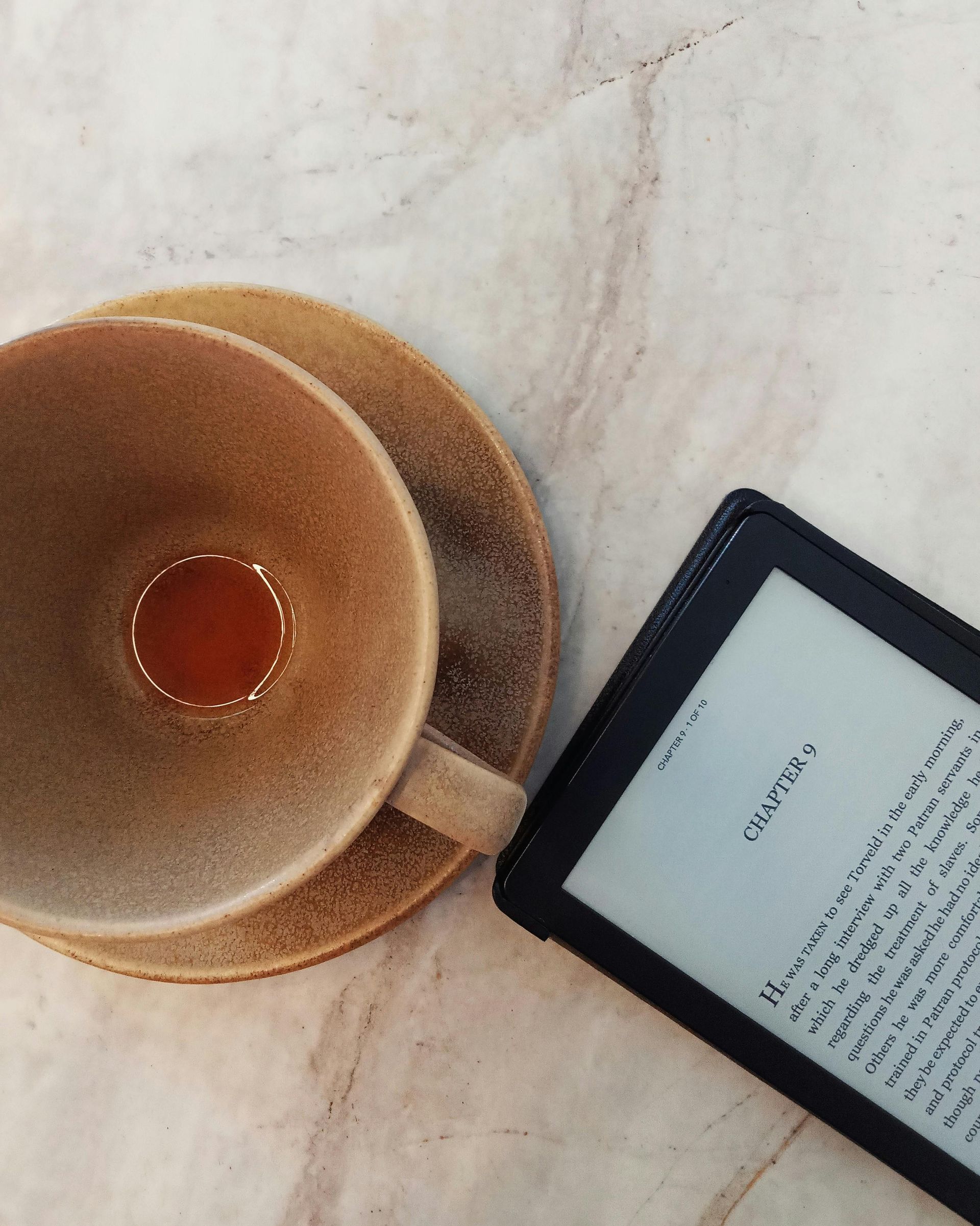 Coffee cup and e-reader on a marble surface. The e-reader displays text.